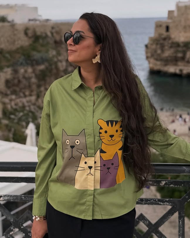 Woman wearing a green shirt with cat designs in an outdoor setting with buildings and ocean.