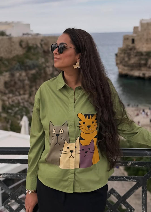Woman wearing a green shirt with cat designs in an outdoor setting with buildings and ocean.
